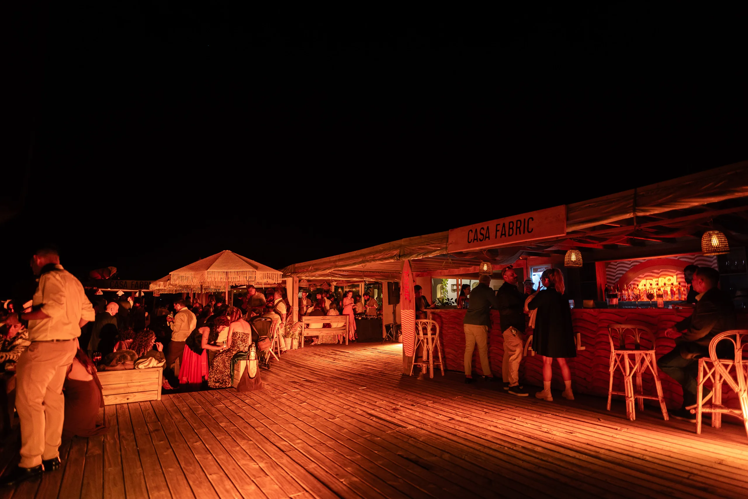 El deck de madera de noche, con los invitados de pie y la cartelería de Casa Fabric iluminada