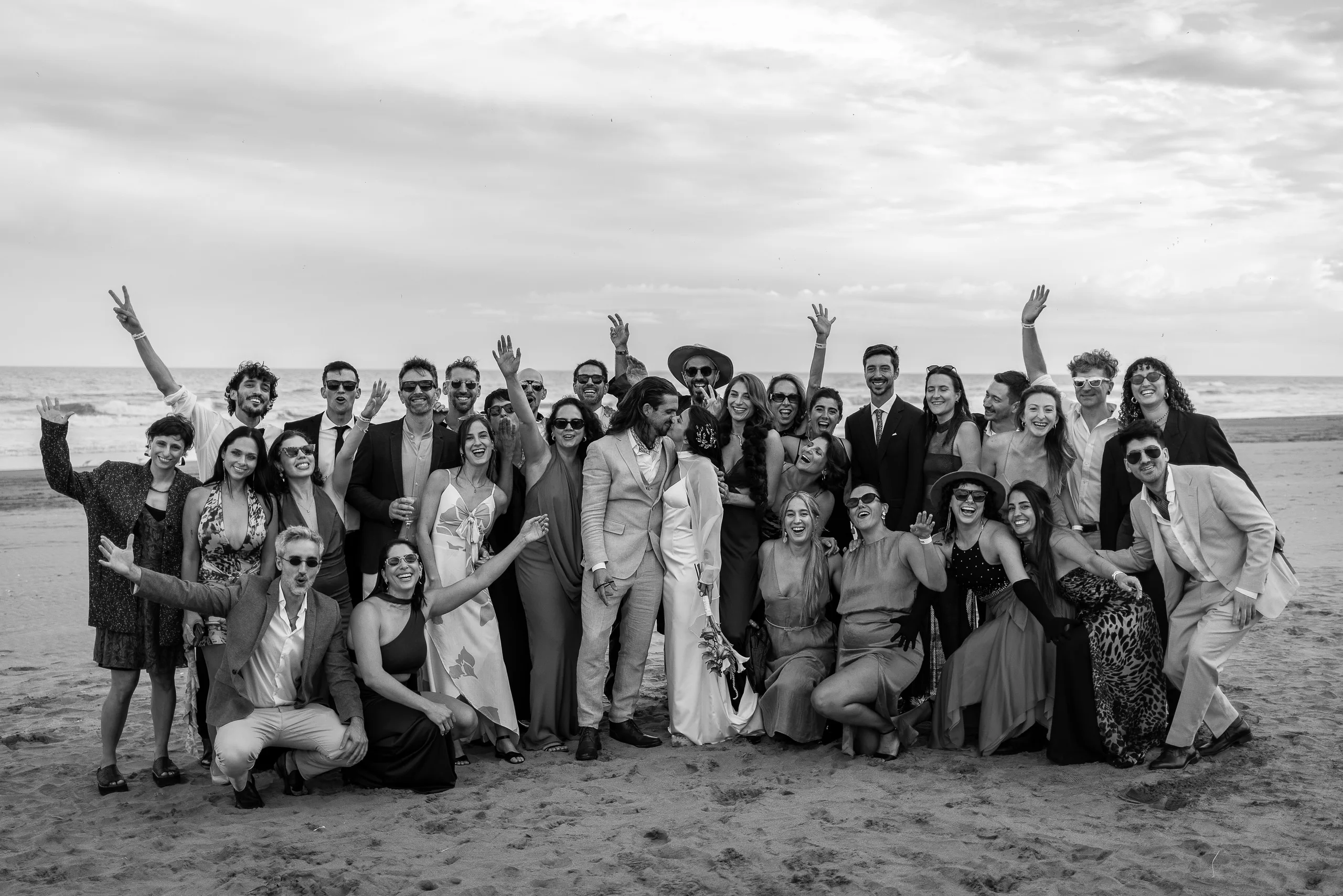 Los novios y todos los invitados con los brazos en alto sobre la arena, con el mar de fondo