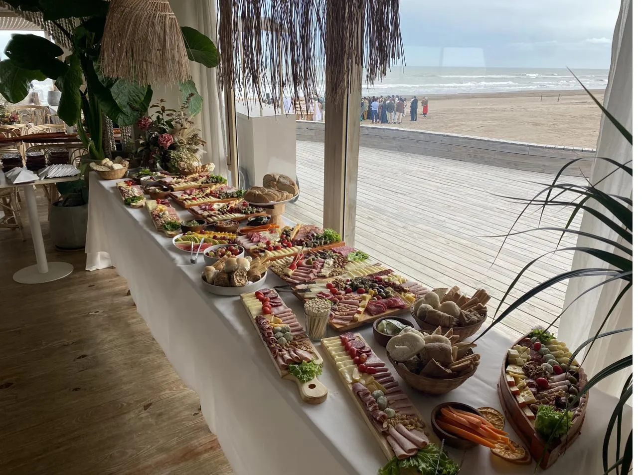 Mesa larga de fiambres, quesos y panes junto al ventanal, con la playa afuera