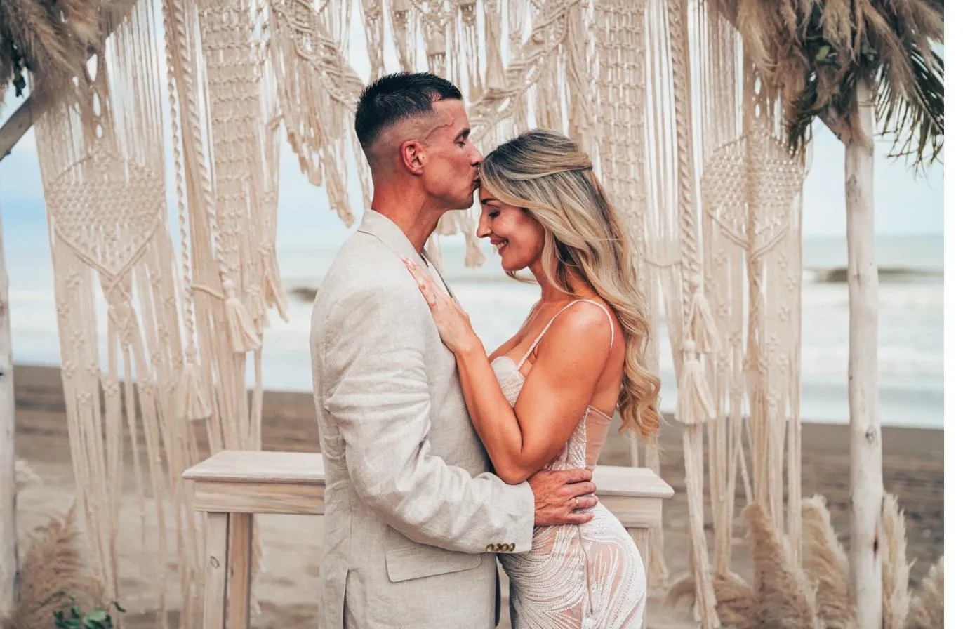 Una pareja abrazada bajo los flecos de macramé, con las palmeras y el mar detrás